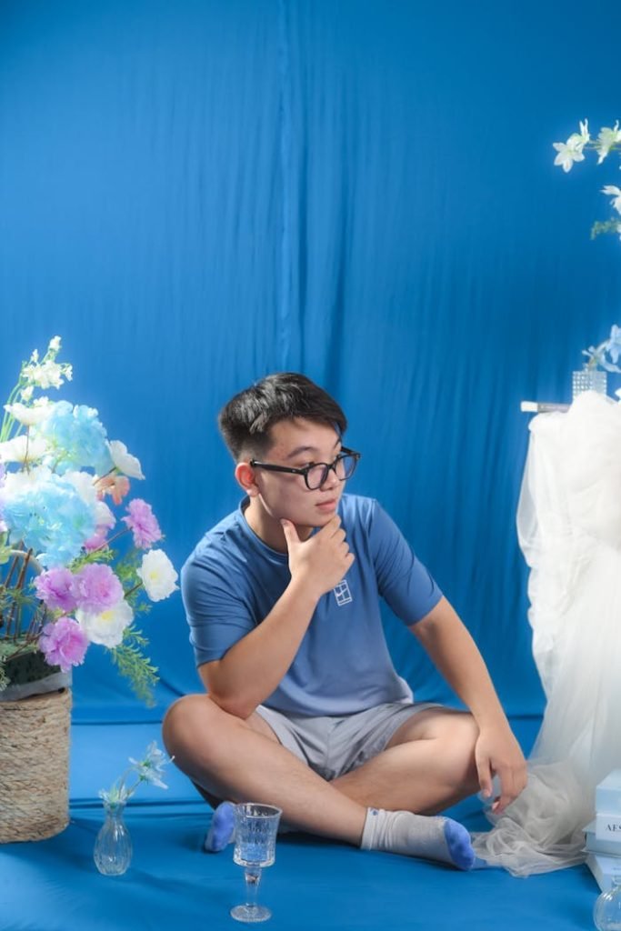 Young man posing thoughtfully in a blue-themed floral studio setup, exuding a calm and pensive atmosphere.